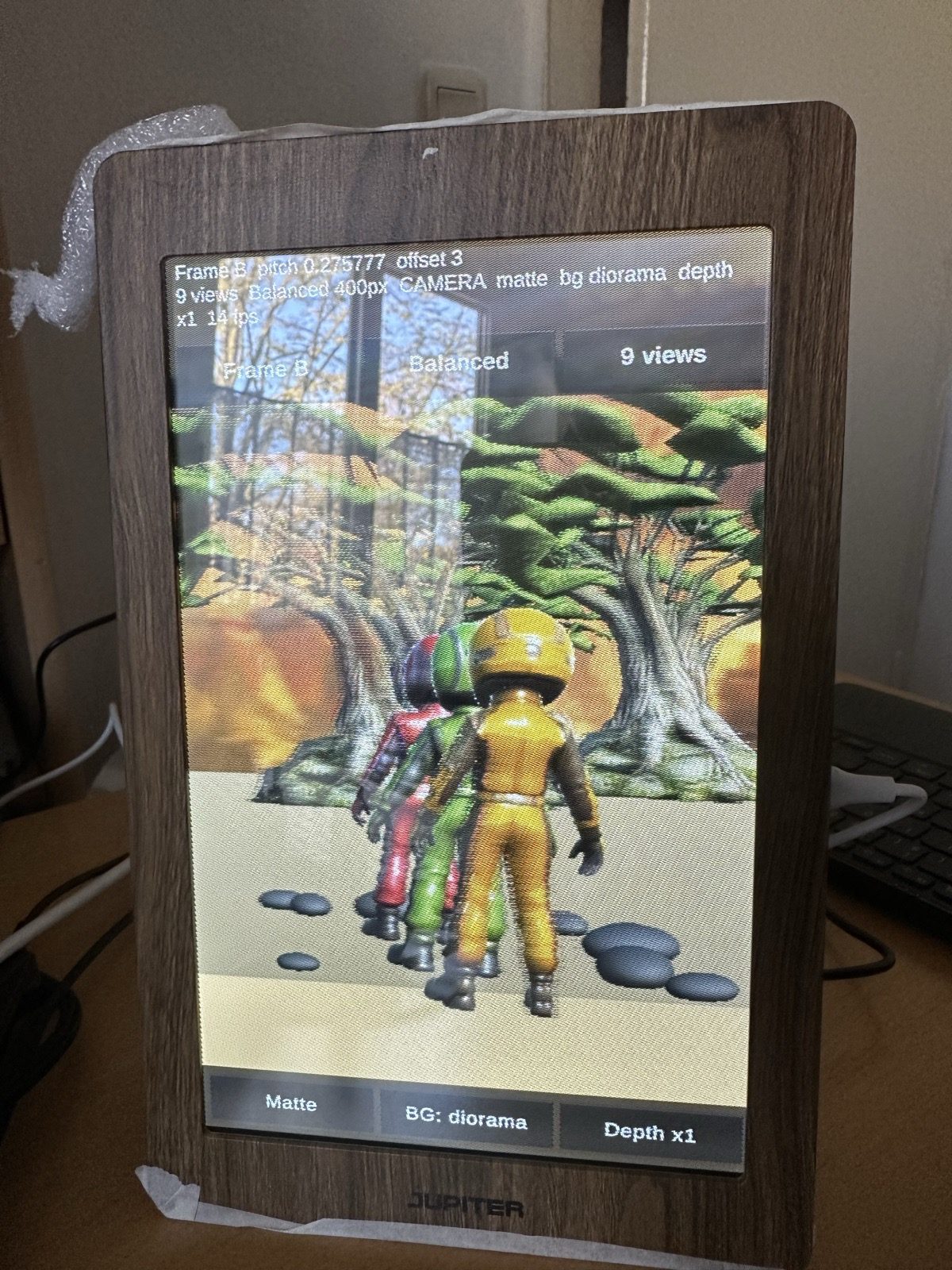 Photo of the JUPITER 3D Frame on a desk, showing three small racing-suit characters walking in front of a painted tree.